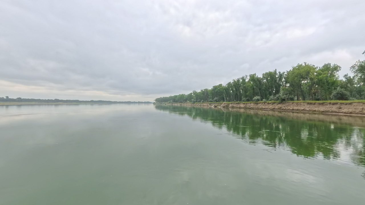 The calm Missouri River reflecting a long wooded bank on the right, 2025