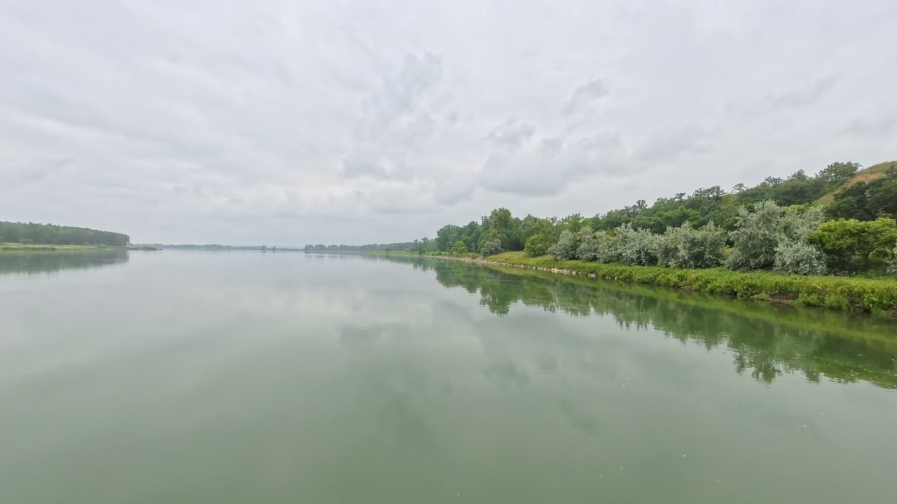 The calm Missouri River beside a leafy bank on the right, under a cloudy sky, 2025