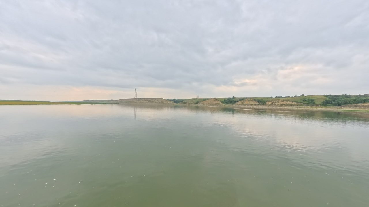 The calm Missouri River below low grassy bluffs under an overcast sky, 2025
