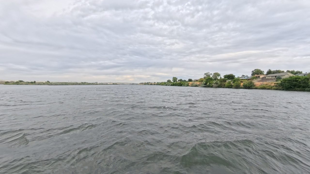A choppy river under a grey, overcast sky, with houses above the right bank and a long bridge in the distance, 2025
