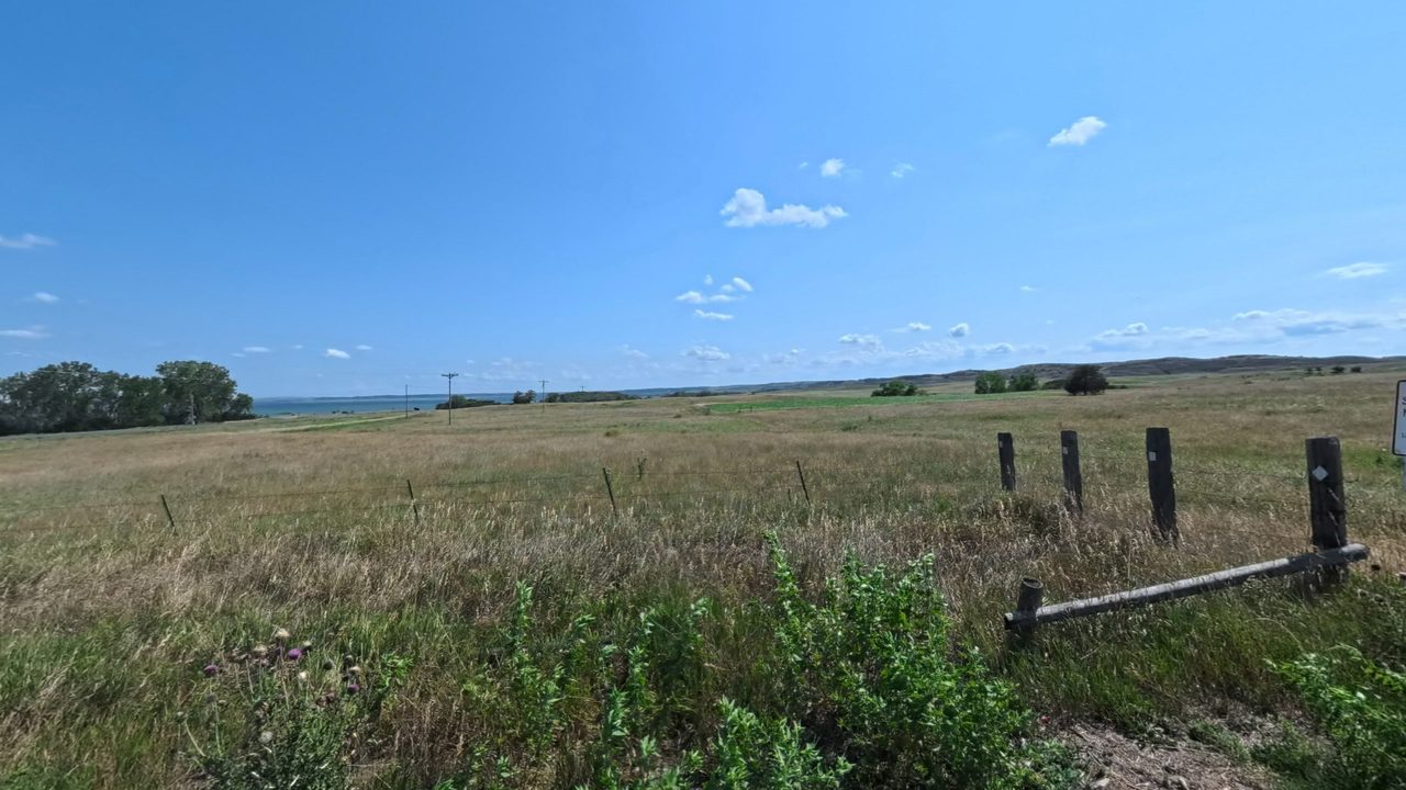 Prairie grassland stretching to the blue water of the Missouri River, with a weathered fence line and a few trees, 2025