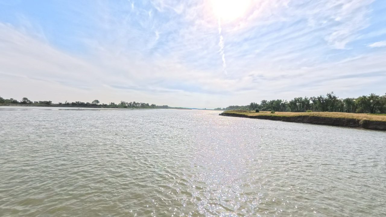 The wide Missouri River glinting under a bright sun, with low wooded banks and streaked clouds, 2025