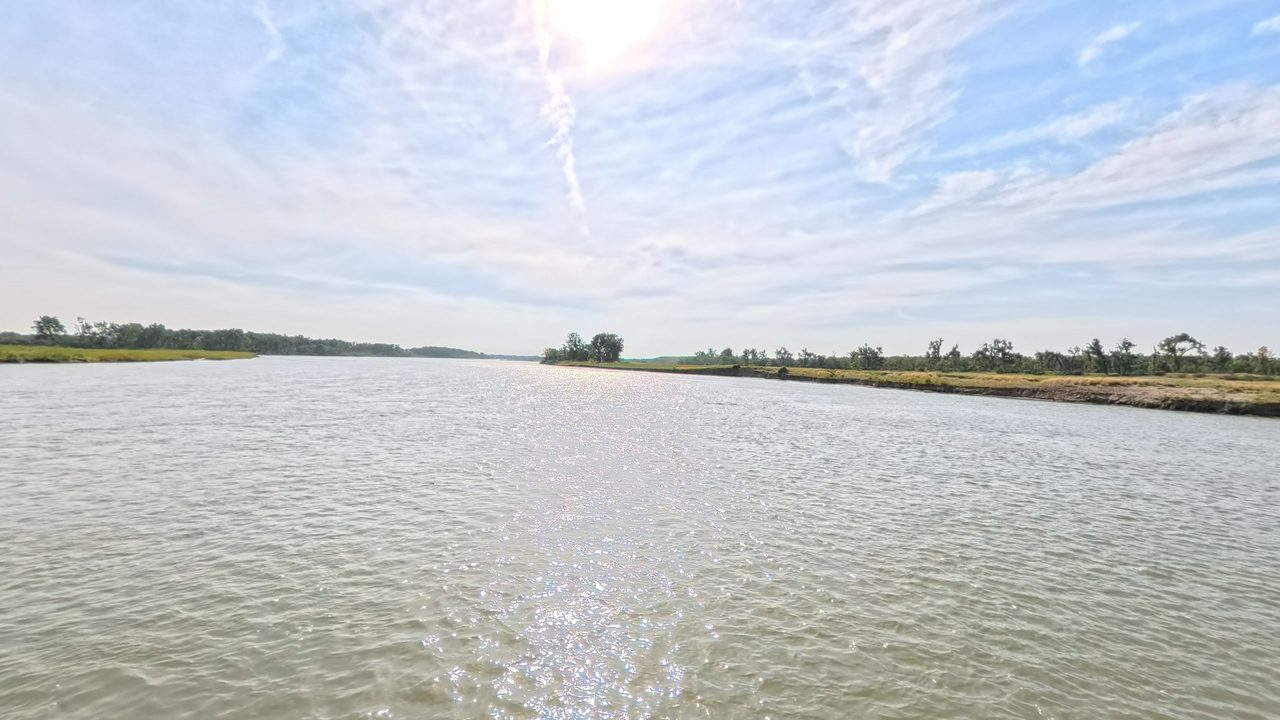 The Missouri River glittering under a high, hazy sun, with low wooded banks on both sides, 2025
