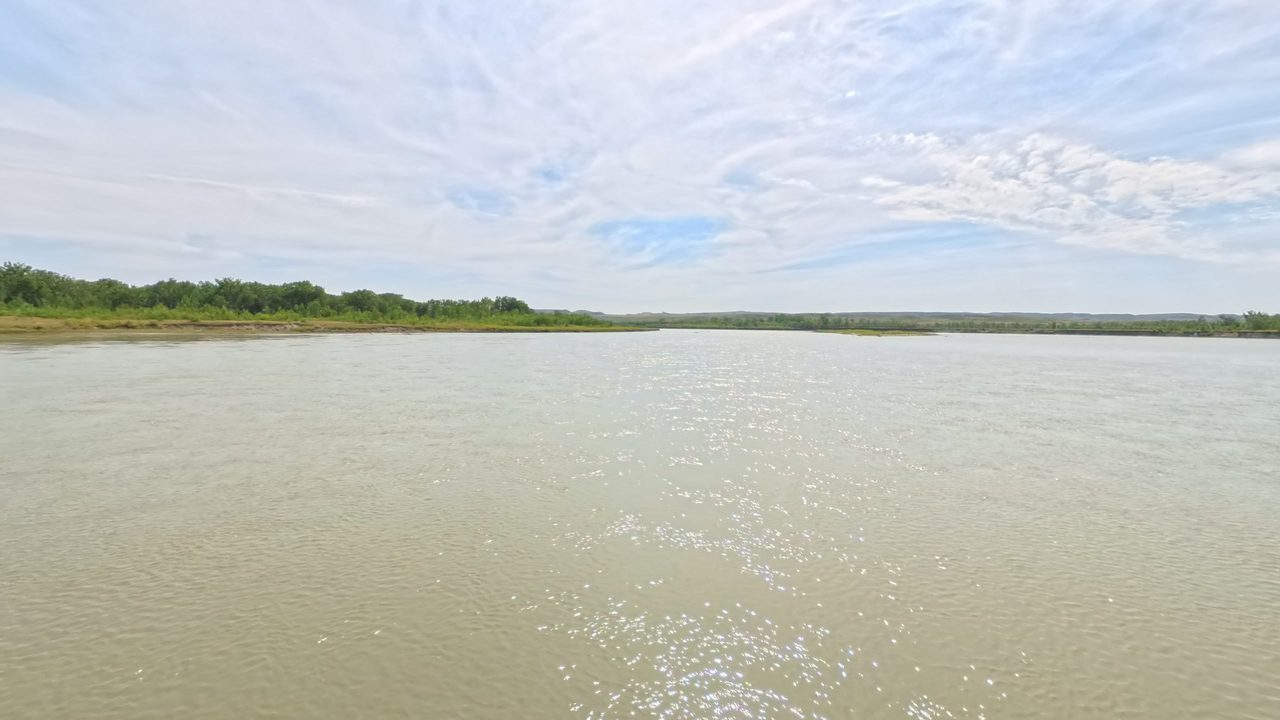 The broad Missouri River under wispy clouds, with a low, wooded bank and distant bluffs, 2025