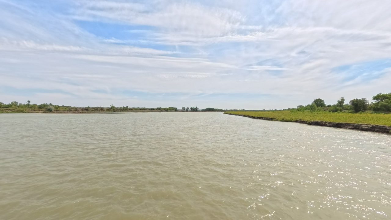The wide Missouri River beside a low grassy bank on the right, under high thin clouds, 2025
