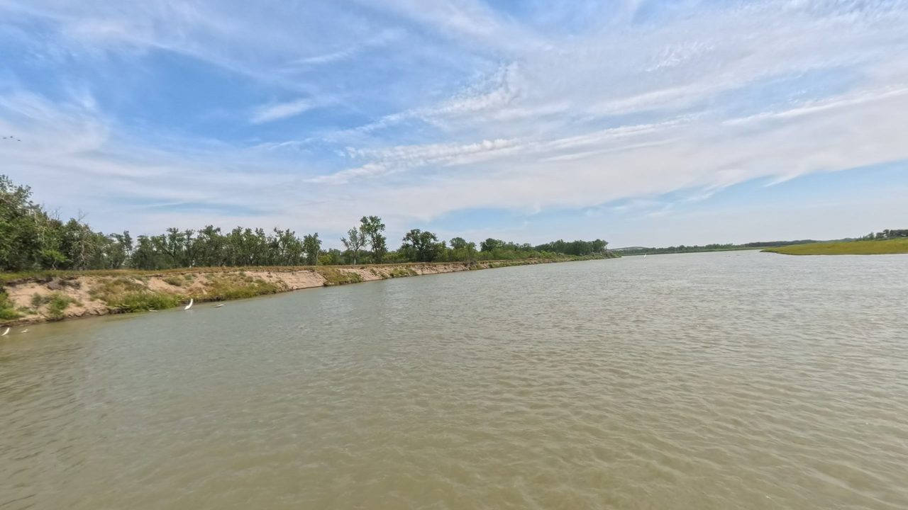 The Missouri River beside a sandy, grassy bank lined with trees on the left, under streaked clouds, 2025