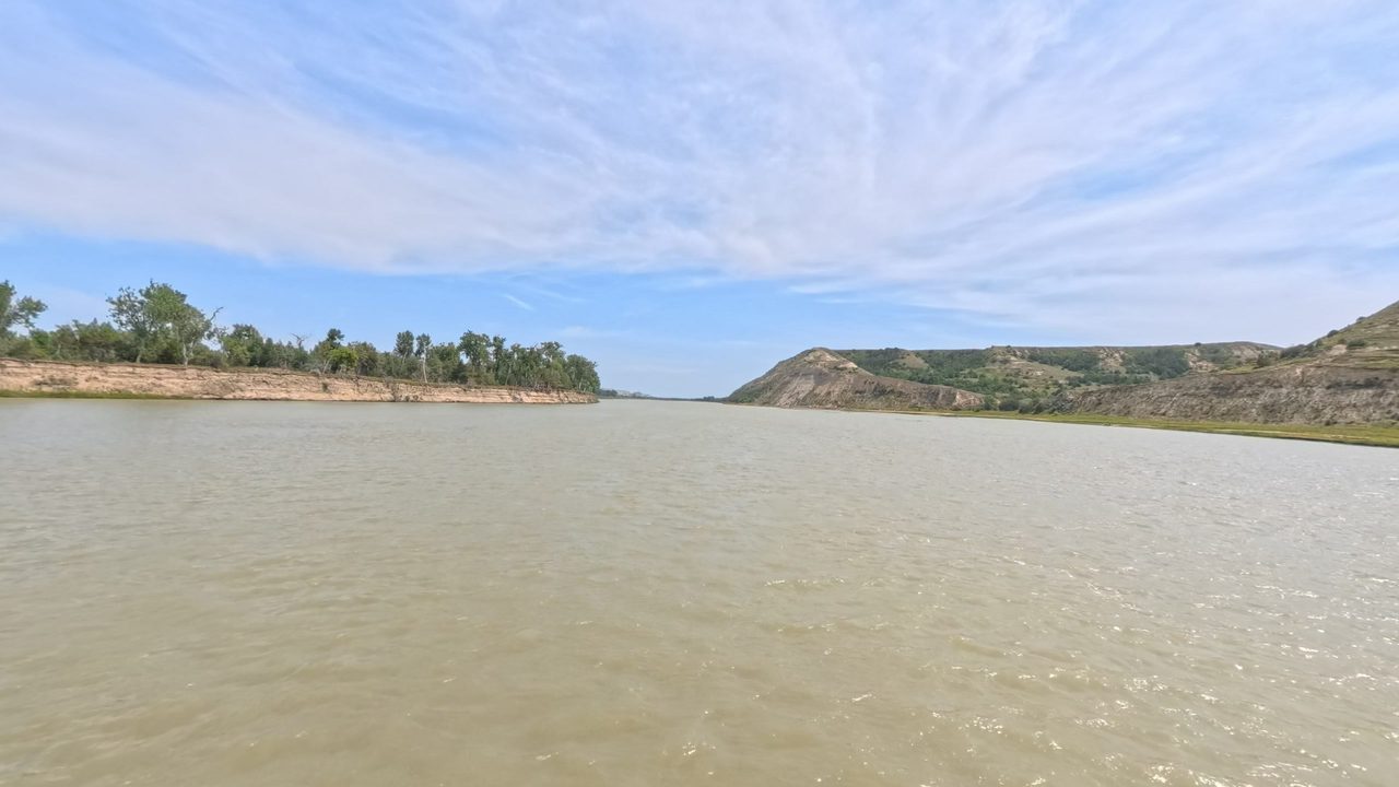 The Missouri River toward a grassy, eroded bluff on the right, with a wooded bank on the left, 2025