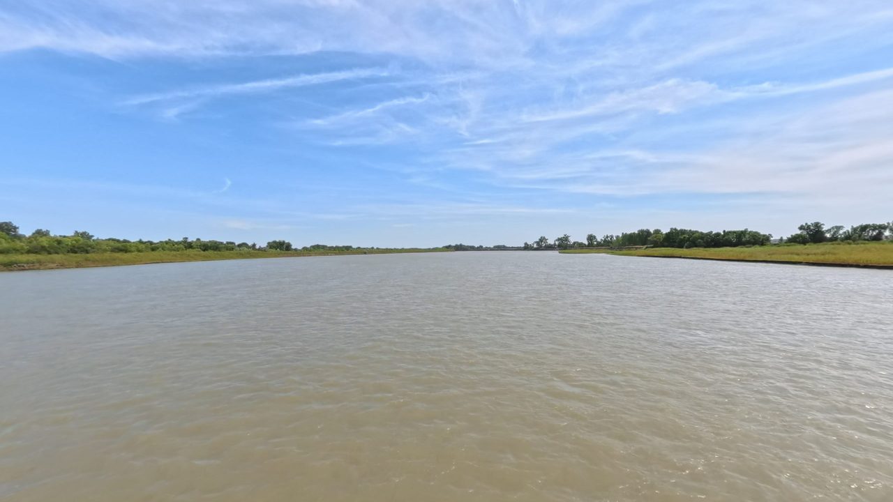 The broad Missouri River between flat, grassy banks under wispy clouds, 2025