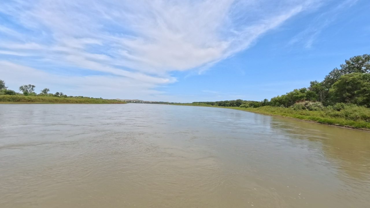 The Missouri River beside a leafy wooded bank on the right, under streaked white clouds, 2025