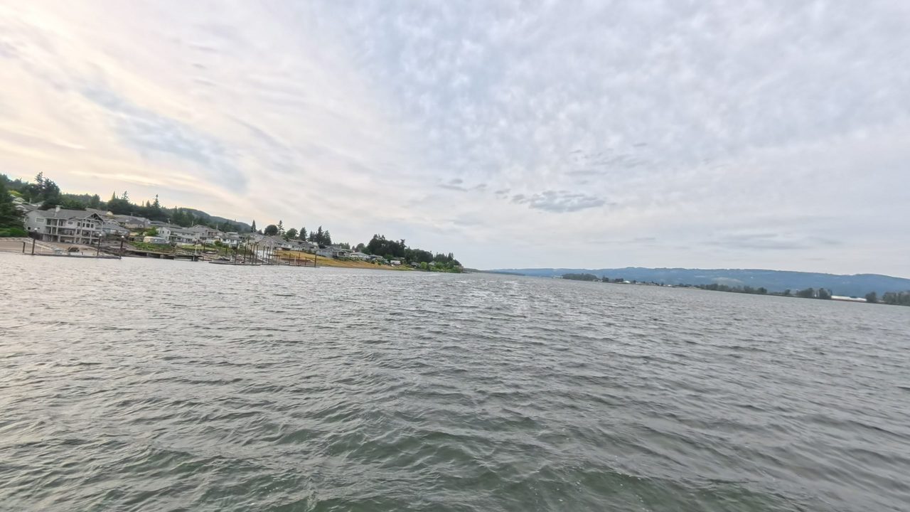 The Columbia River under a mottled grey sky, with waterfront houses and docks on the left shore, 2025