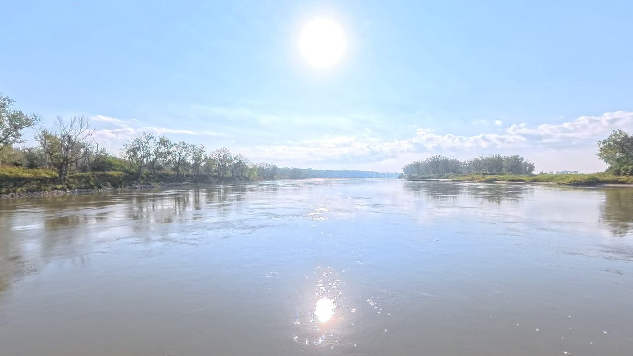 The Missouri River under a high sun, with light glittering on the water and trees on both banks, 2025