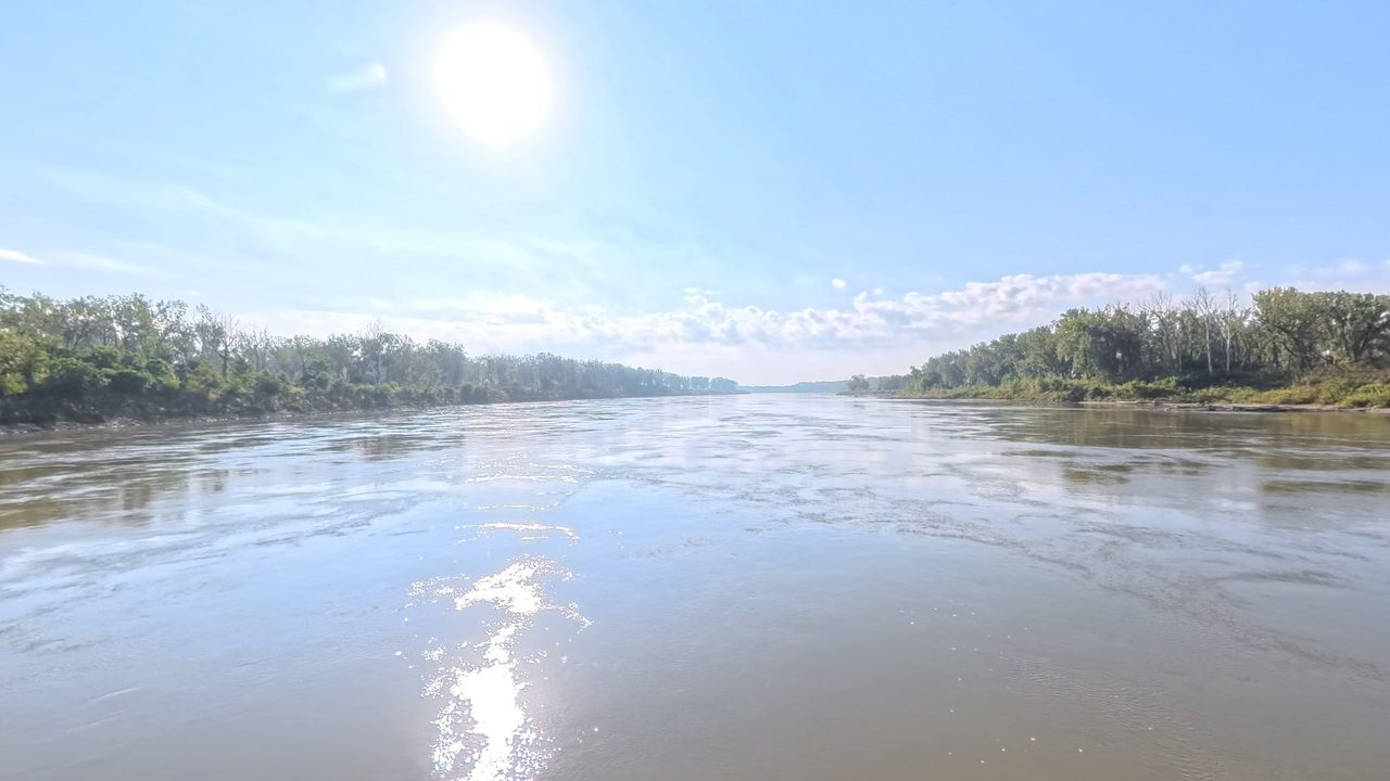 The Missouri River between wooded banks under a bright sun and scattered clouds, 2025