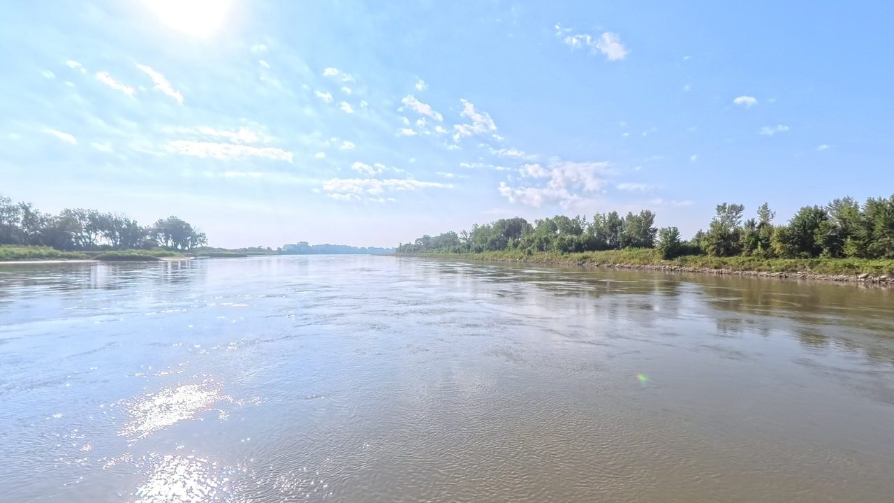 The Missouri River between leafy banks under a blue sky with small white clouds, 2025