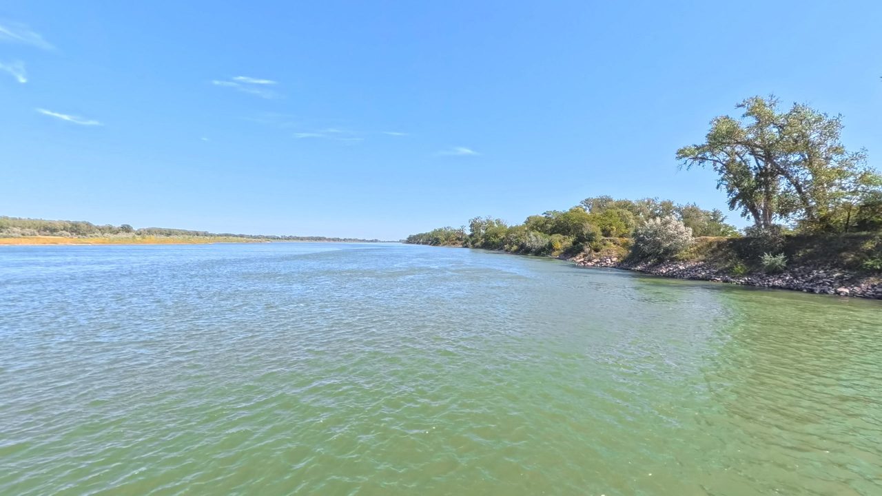 The green Missouri River beside a rocky, tree-lined bank under a clear sky, 2025