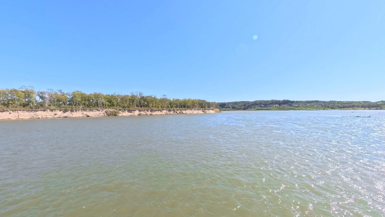 The Missouri River below a sandy bank lined with trees, under a clear sky, 2025