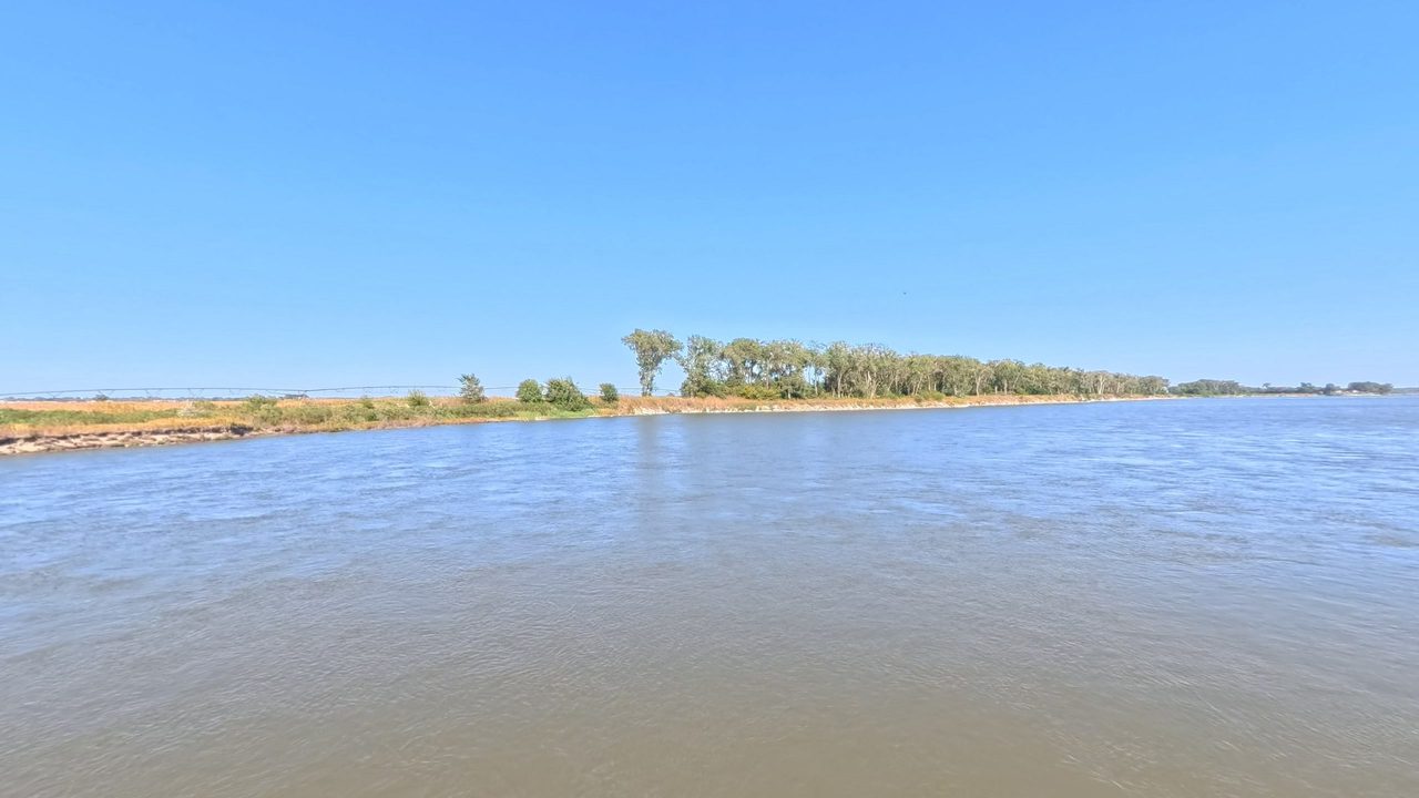 The Missouri River around a long, sandy island lined with trees, under a clear sky, 2025