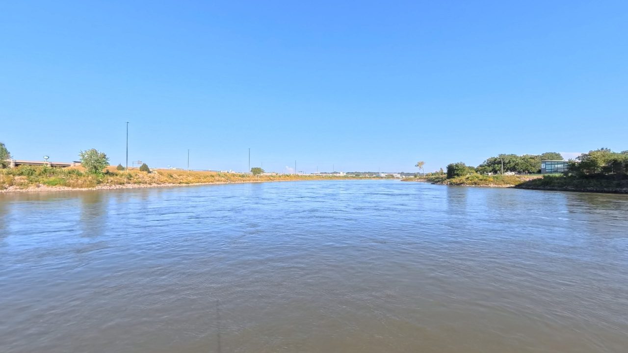 The Missouri River under a clear blue sky, with grassy banks and a riverside building, 2025