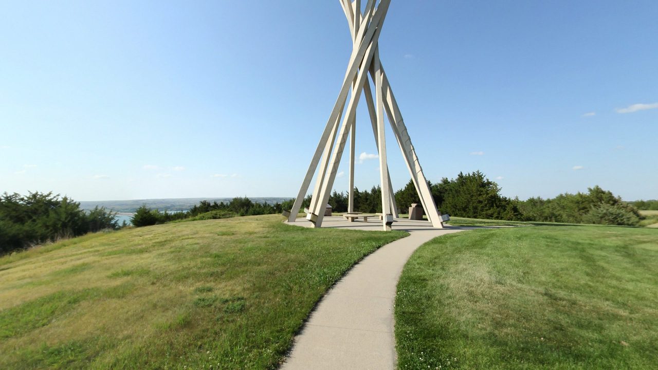 A paved path climbing a grassy hilltop to a tall tipi-shaped sculpture, with the Missouri River glimpsed beyond, 2025