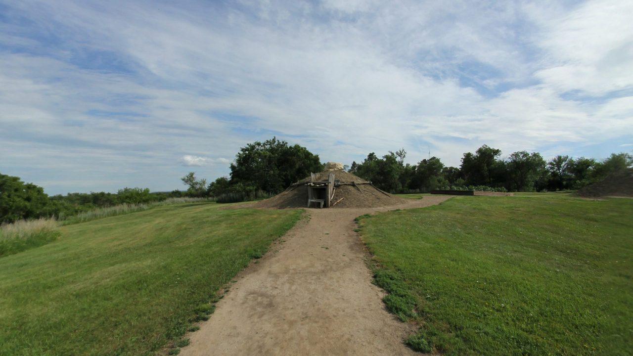 A dirt path across mown grass toward a domed earth lodge among trees, 2025