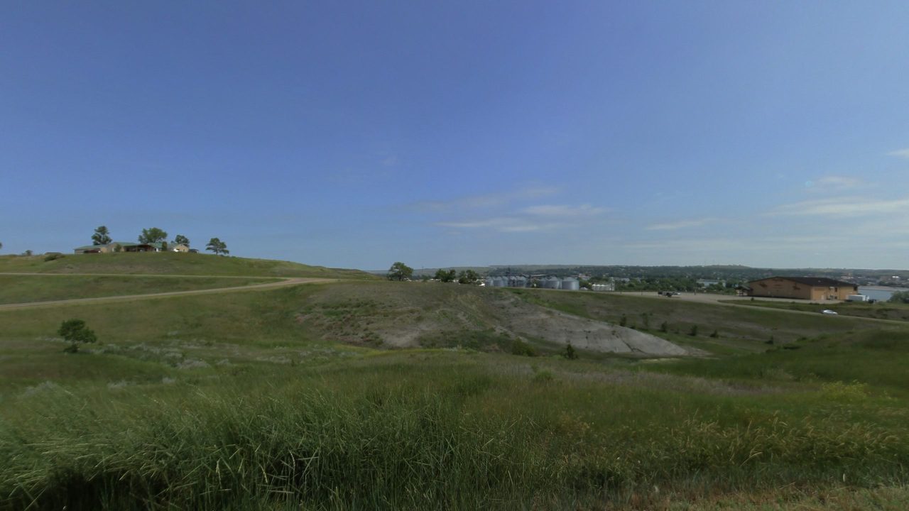 Grassy hillsides and an eroded slope, with houses and grain bins beyond, 2025