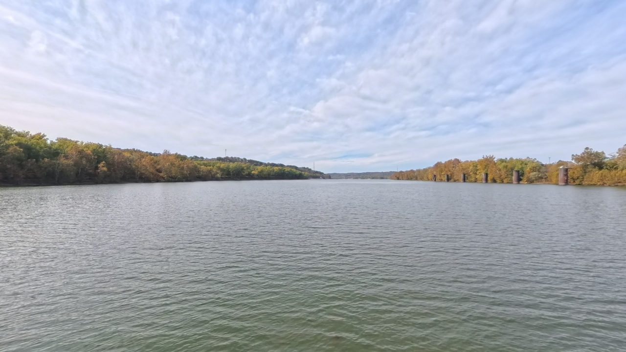 A wide river between autumn-tinged wooded banks under rippled clouds, with old bridge piers on the right, 2025