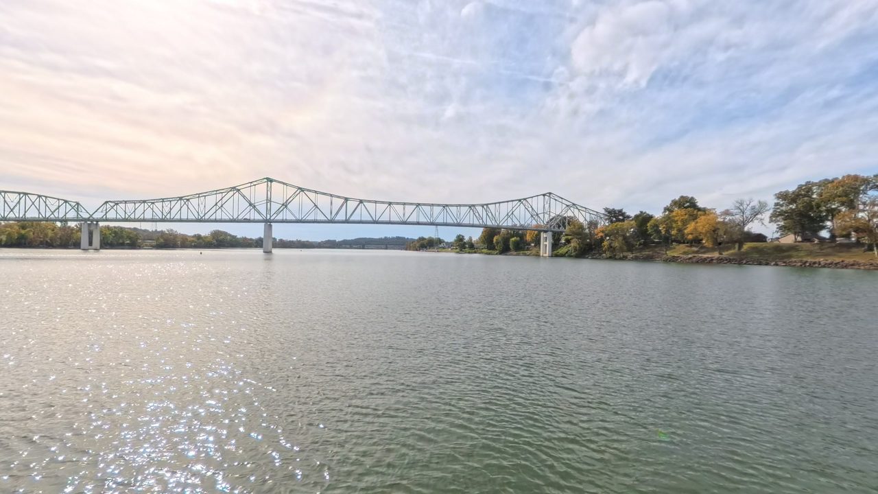 The Ohio River below a long steel truss bridge, with autumn trees along the right bank and sun glinting on the water, 2025