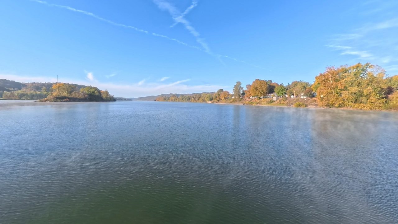 The calm Ohio River under a blue sky, with houses among trees in autumn color along the right bank, 2025