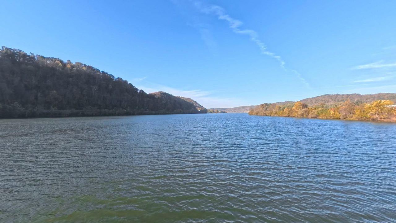 The Ohio River below a steep wooded hillside on the left, with autumn trees on the right, 2025