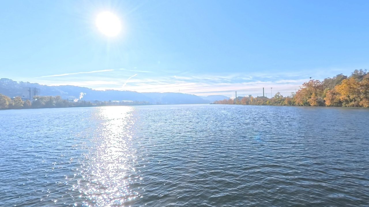 The Ohio River glittering under a bright sun, with autumn trees on the right bank and stacks in the distance, 2025