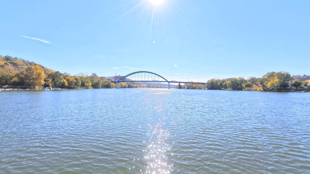 The Ohio River toward an arched bridge, with autumn woods on both banks under a bright sun, 2025