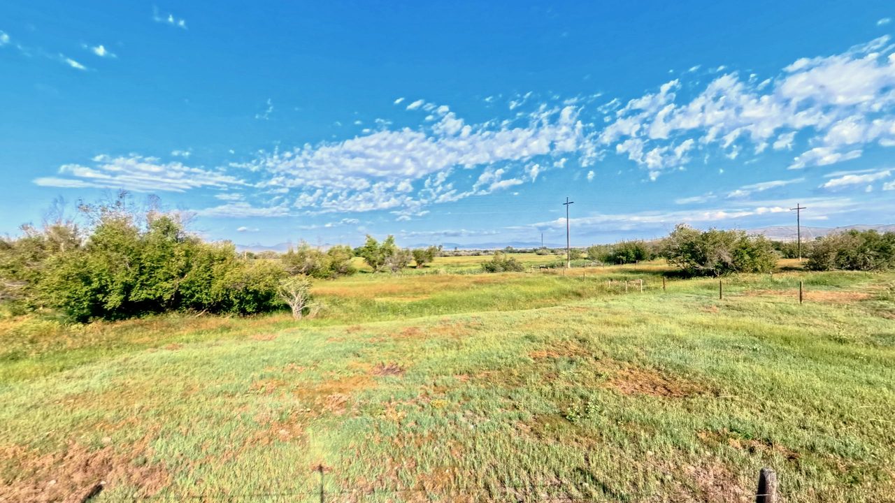 A green pasture with shrubs and fence posts below distant mountains, under scattered clouds, 2026