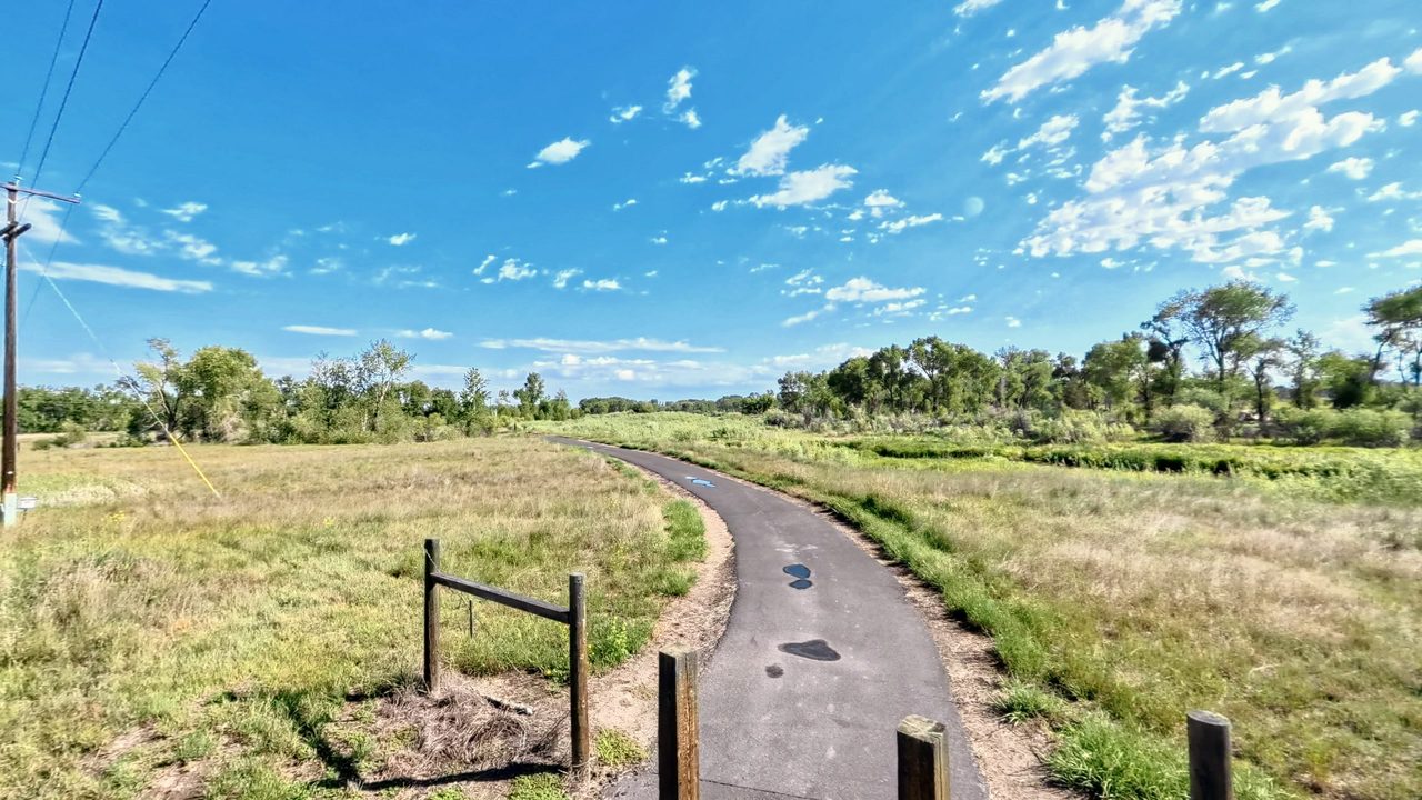 A paved trail curving through green pasture toward a line of cottonwoods, with wooden posts in the foreground under a blue sky with scattered clouds, 2026