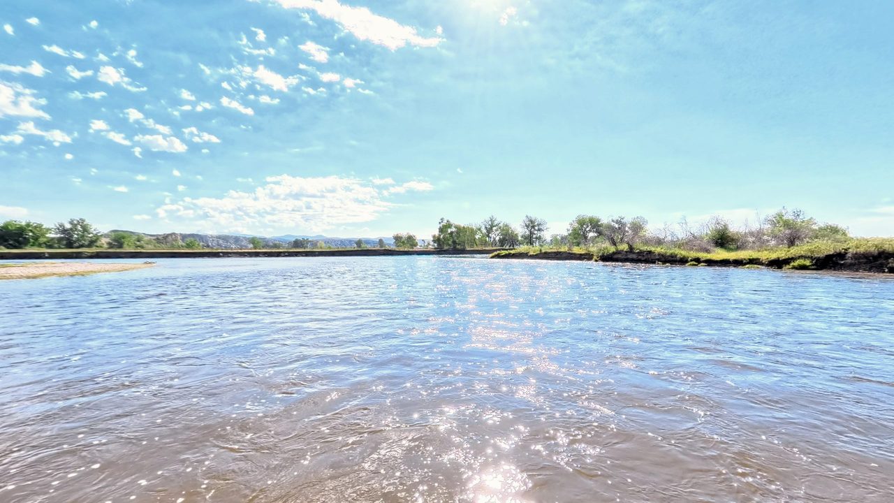A wide, rippling river glittering under scattered clouds, with low trees along both banks, 2026