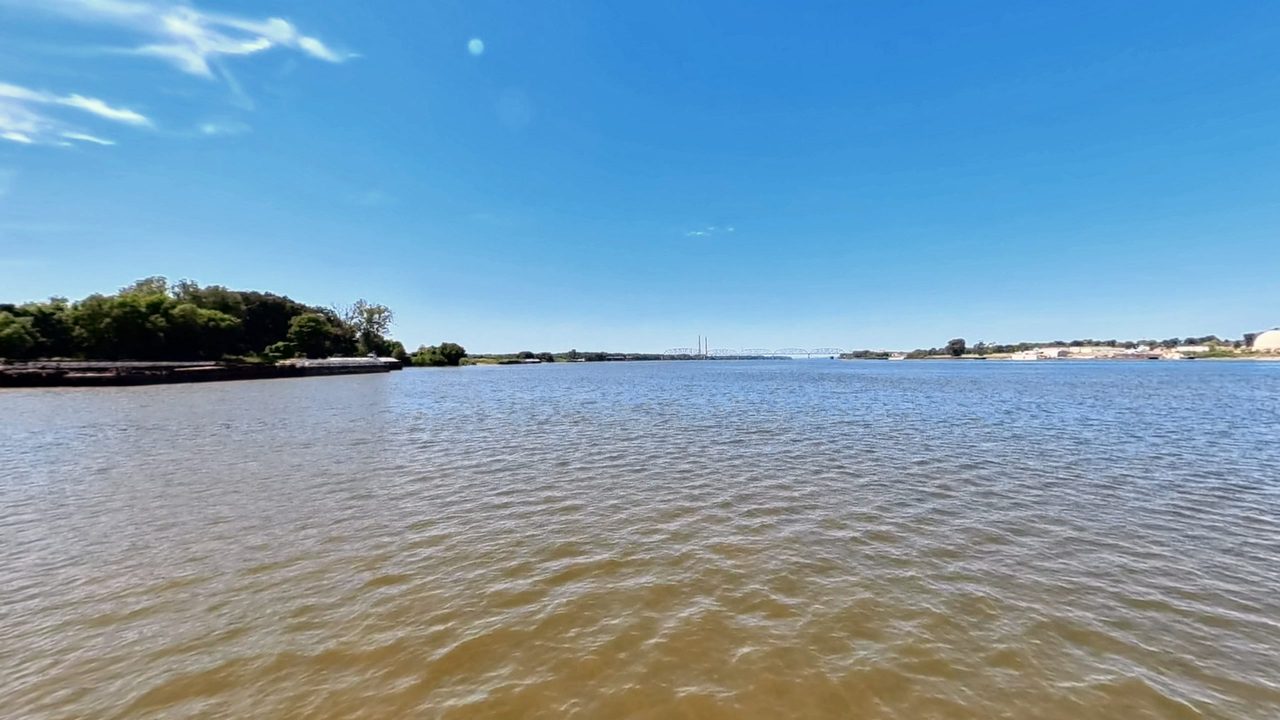 The Ohio River with a wooded point and moored barges on the left, under a clear blue sky, 2026