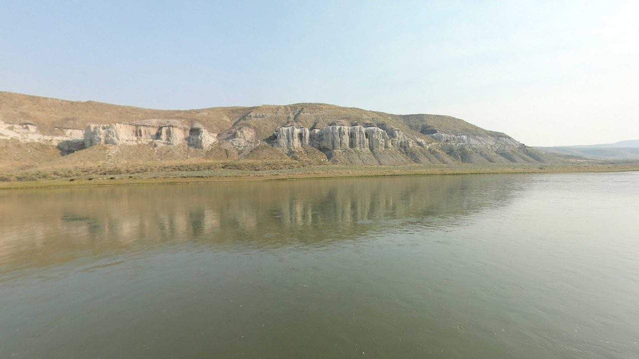 White sandstone cliffs above the calm Missouri River, with their reflection in the water under a hazy sky, 2021