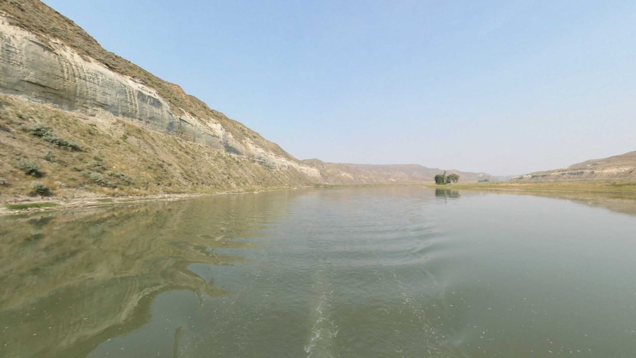 The calm Missouri River between dry, eroded hills, with a small cluster of trees on a far point, 2021
