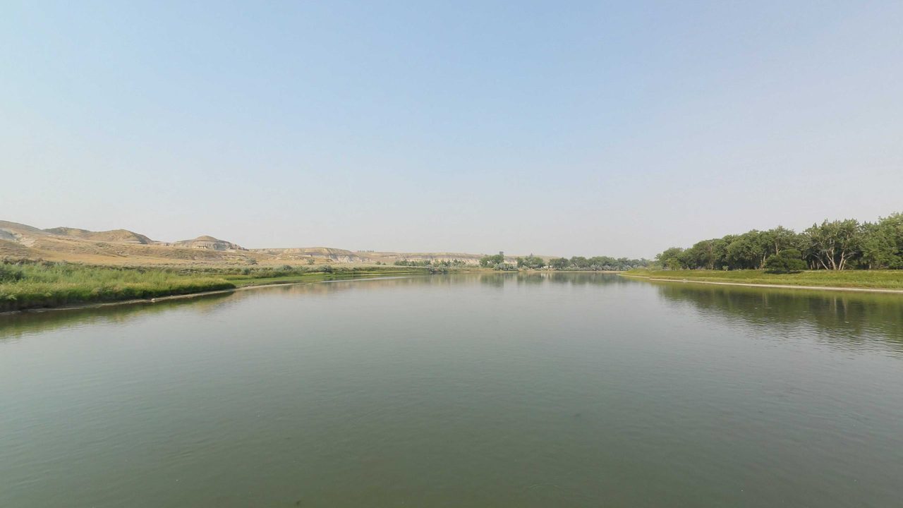The Missouri River between grassy banks, with cottonwoods on the right and low hills beyond, 2021