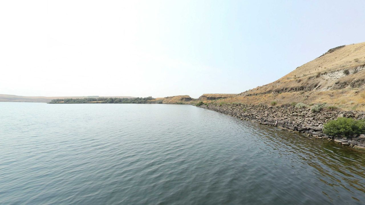 The calm Snake River below a rock embankment and a dry grassy hill on the right, under a hazy sky, 2021