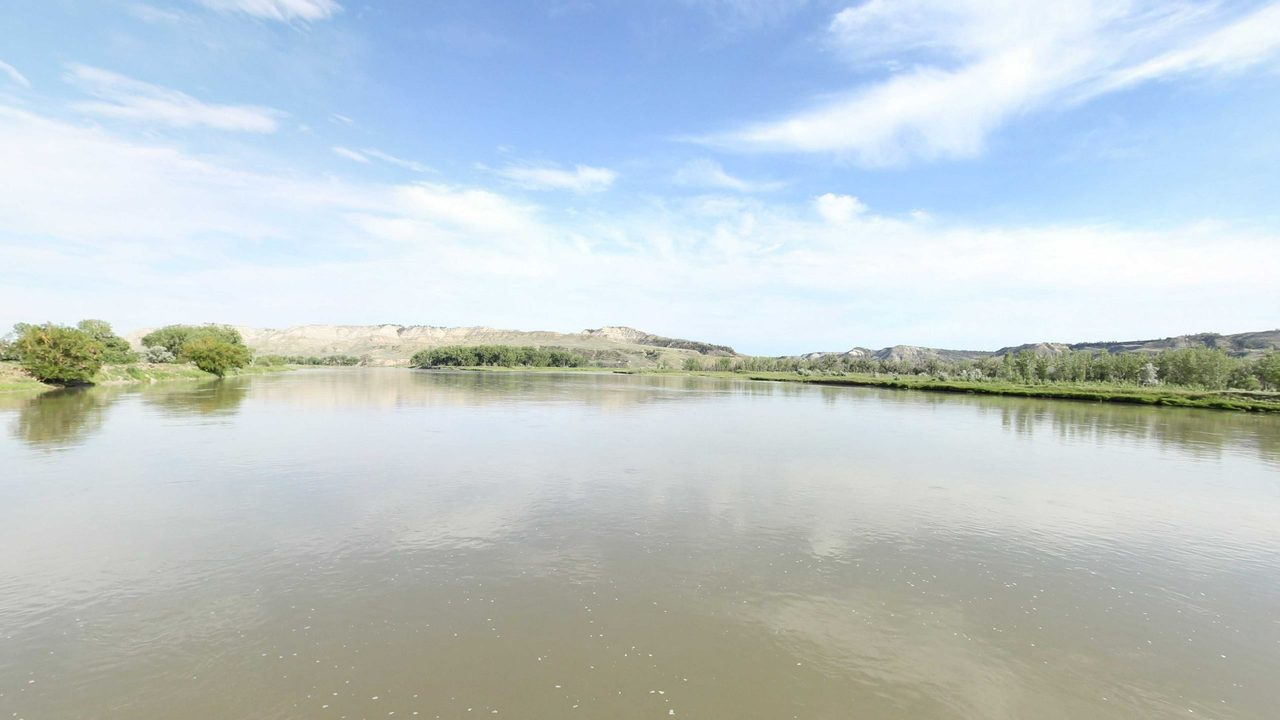 The calm Missouri River between low banks with cottonwoods, below long, pale bluffs, 2022
