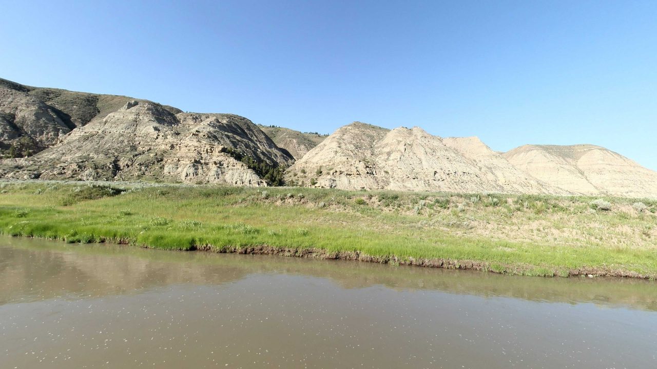 Rugged pale badland cliffs above a green, grassy riverbank, 2022