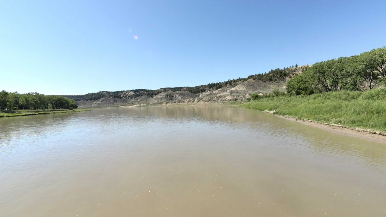 The Missouri River below pale badland bluffs, with a grassy bank and trees on the right, 2022
