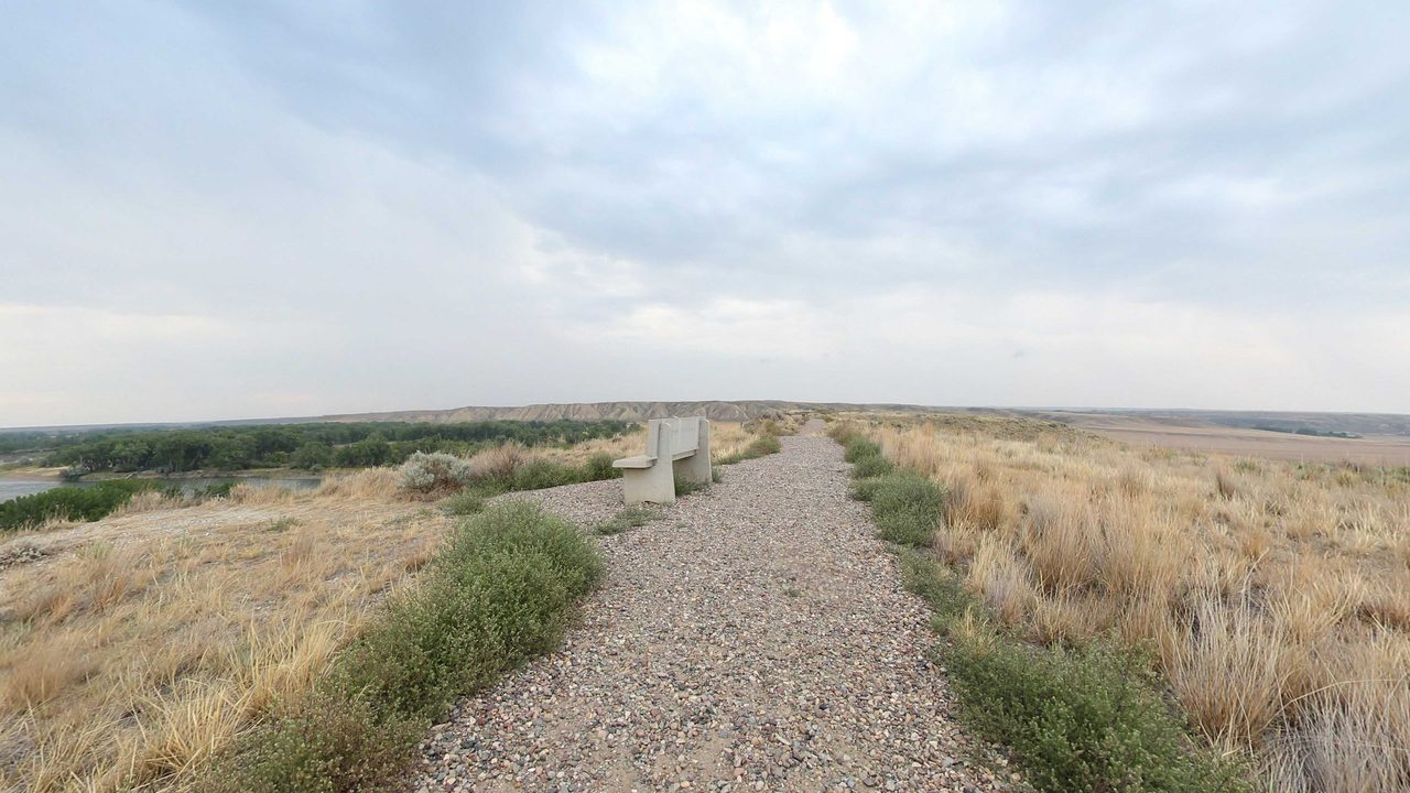 A gravel path with a concrete bench along a grassy bluff, above a river and its wooded bottomland, 2022