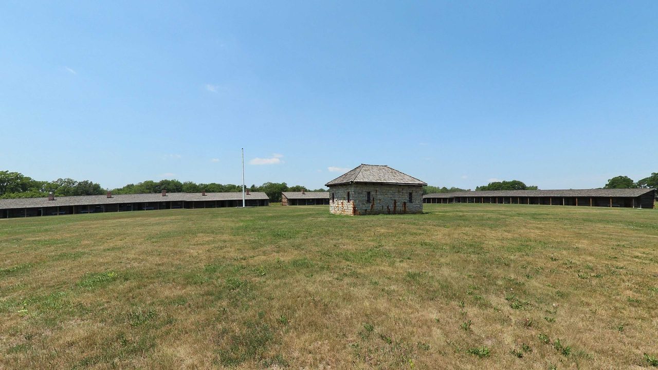 A mown lawn with a stone blockhouse, long log barracks and a tall flagpole under a blue sky, 2023