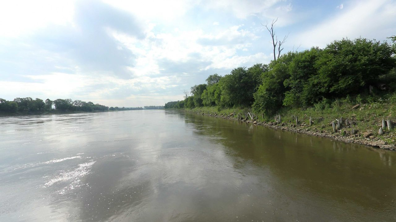 The Missouri River beside a wooded bank lined with rocks, under scattered clouds, 2023
