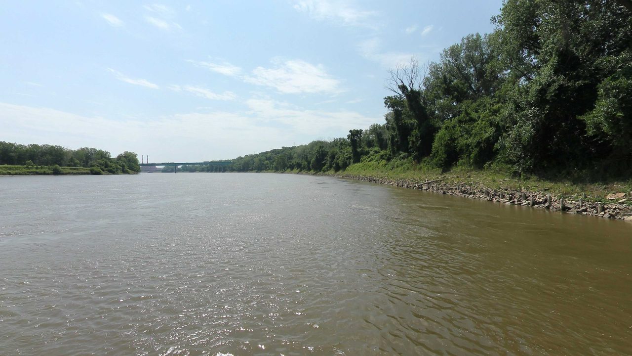The Missouri River beside a wooded bank lined with rocks, with a bridge in the distance, 2023