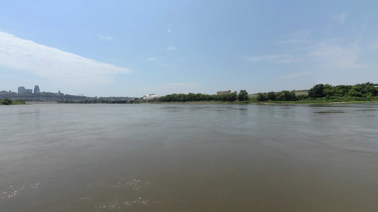 The Missouri River toward a distant city skyline, with a low wooded bank on the right, 2023