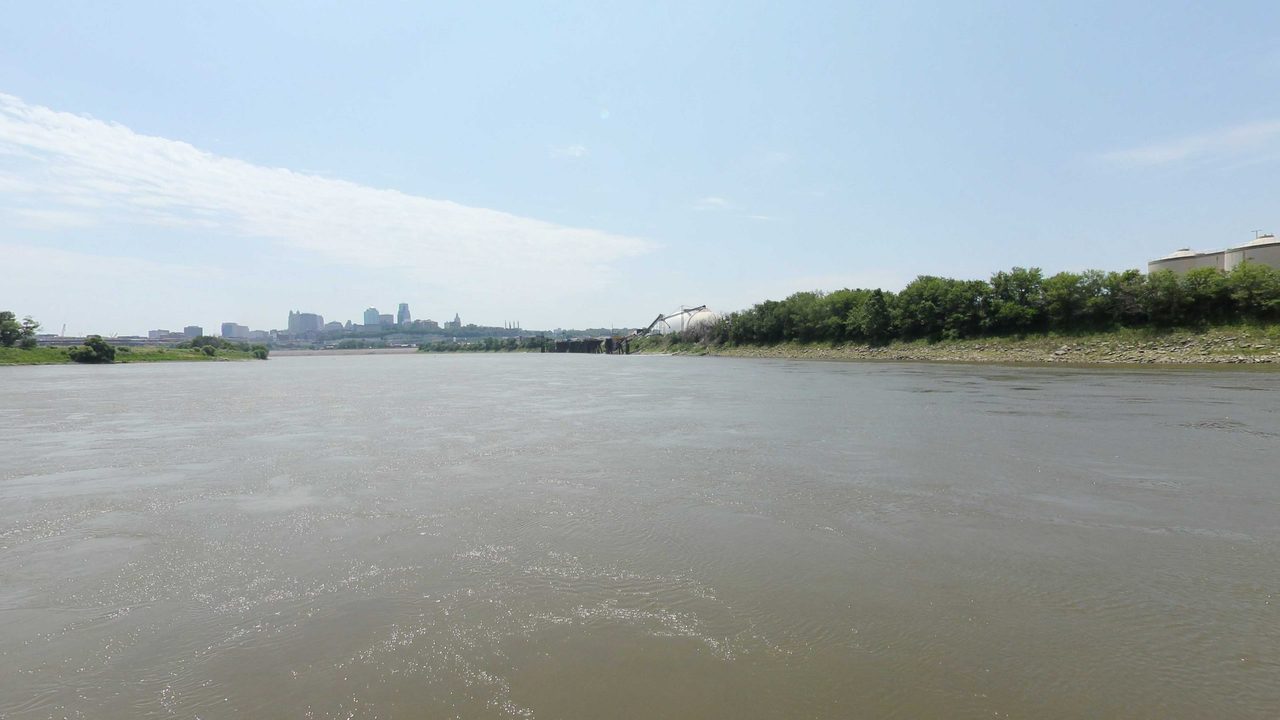 The Missouri River with a distant city skyline and storage tanks above the wooded right bank, 2023