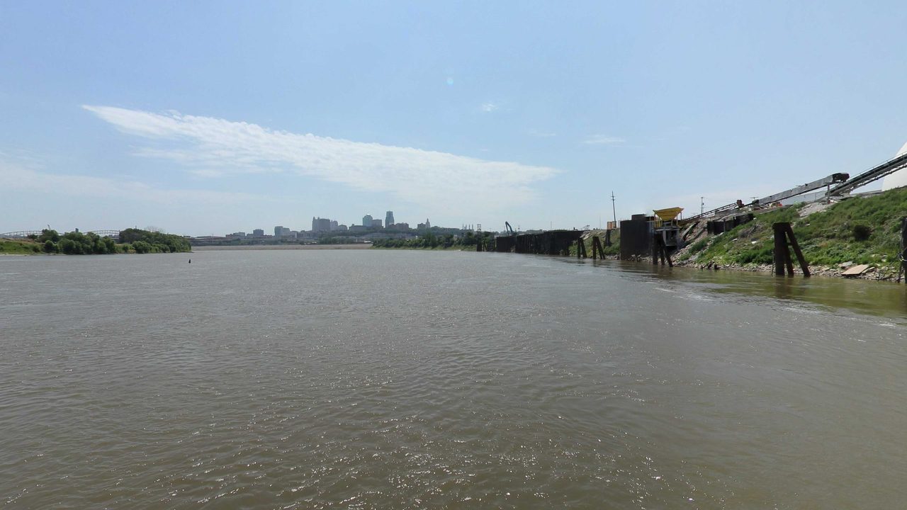 The Missouri River toward a distant city skyline, with an industrial dock and pilings on the right bank, 2023