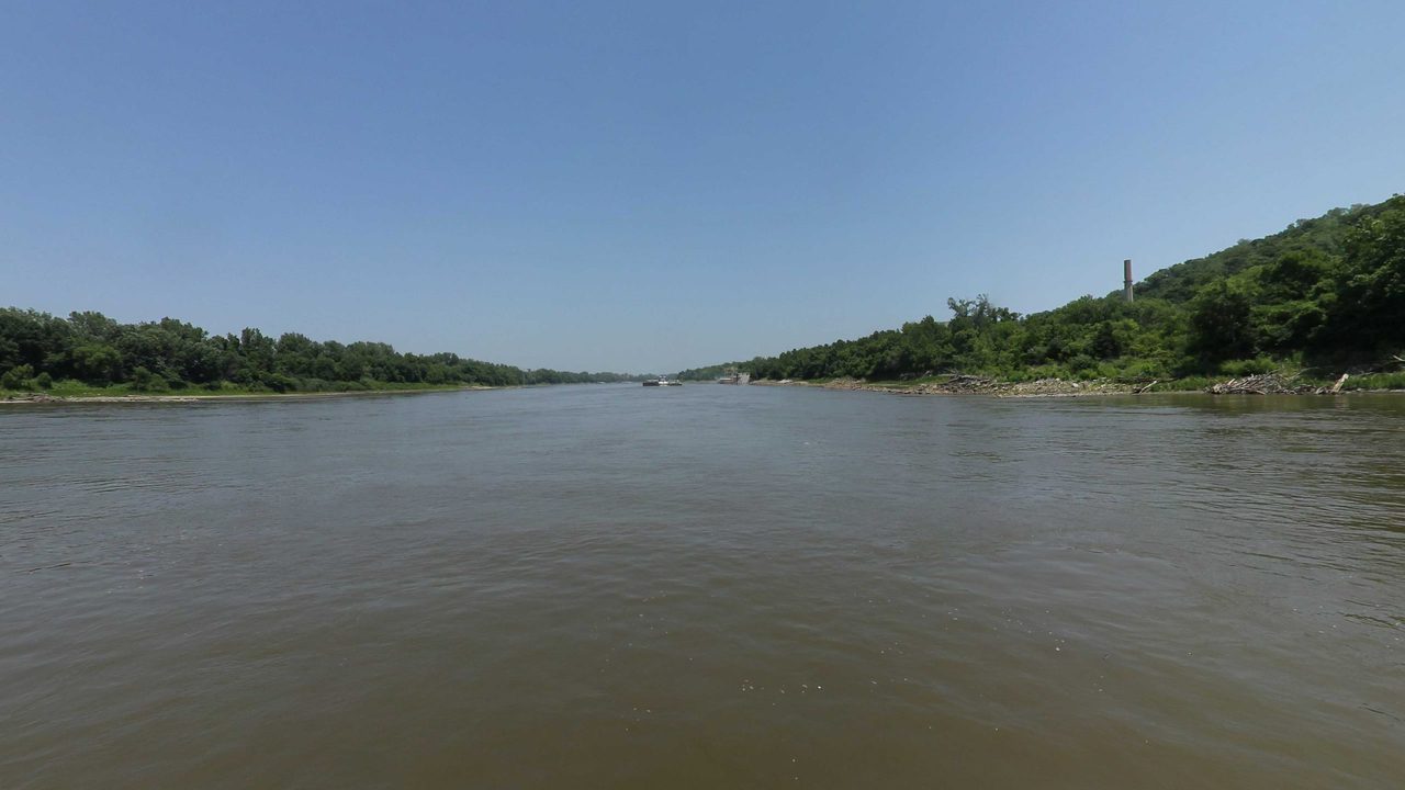 The Missouri River between wooded banks, with a smokestack on the hill to the right, 2023