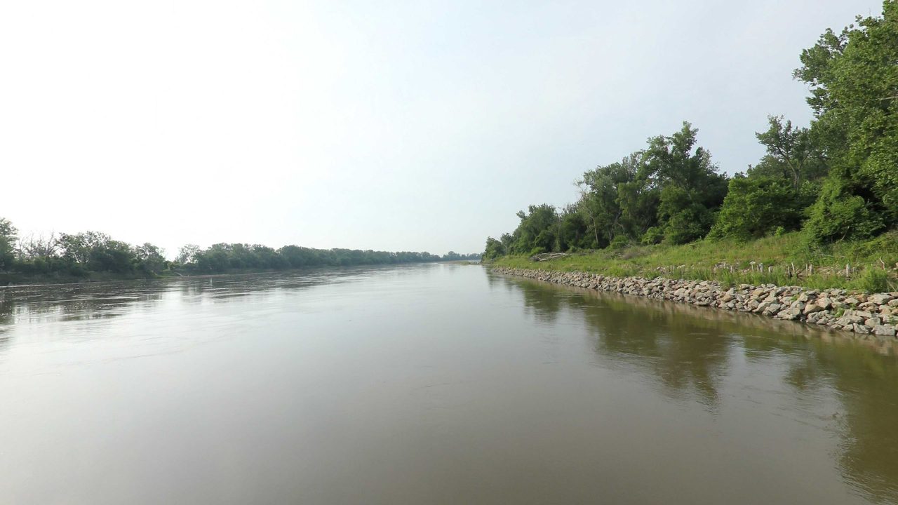 The Missouri River beside a rock-lined bank below leafy trees, under a pale sky, 2023